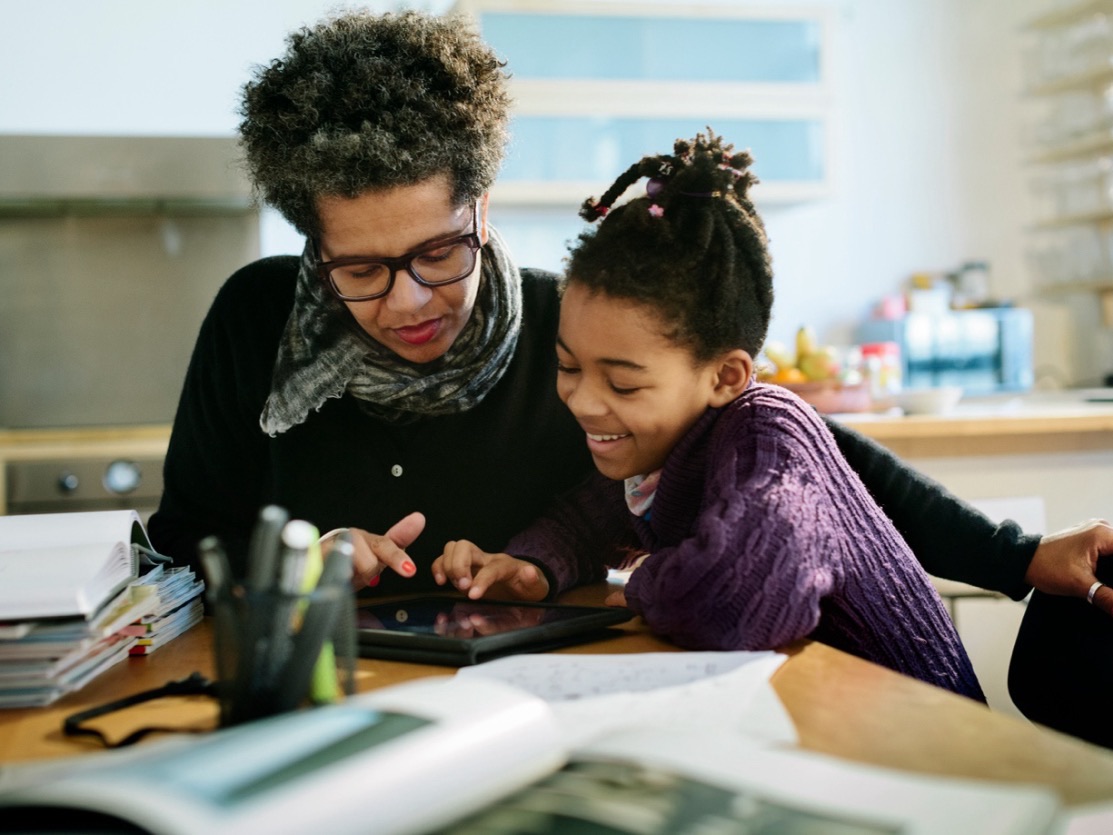 A woman sits beside a smiling girl at a table, helping her with a tablet