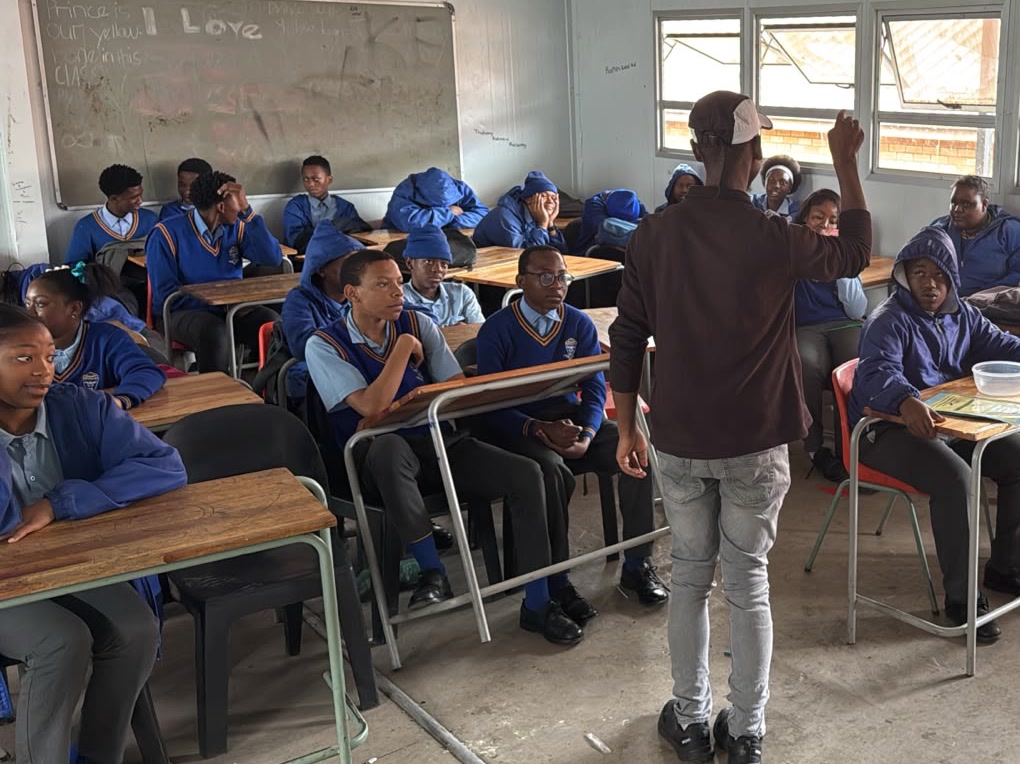 A facilitator standing to speak with a classroom of learners in school jumpers, seated at wooden desks